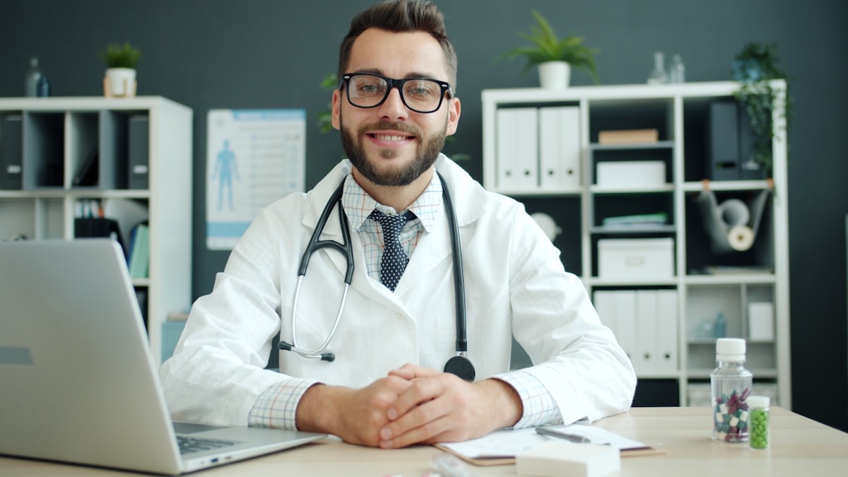 A GP wearing a stethoscope sitting at a consulting-room desk with a laptop