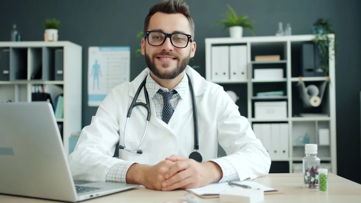 A GP wearing a stethoscope sitting at a consulting-room desk with a laptop